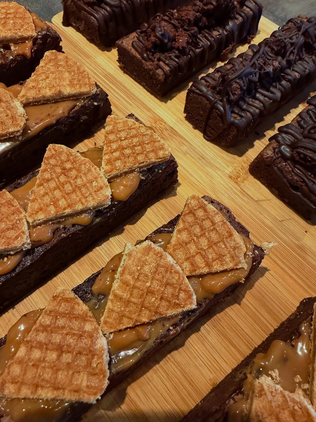Trays of brownies on a wooden board, some topped with caramel and wedges of waffle biscuit, others with chocolate drizzle