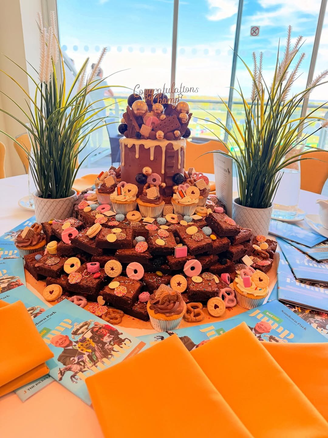 A tall celebration cake covered in biscuits and sweets, surrounded by brownies and cupcakes on a table by a big window at a racecourse