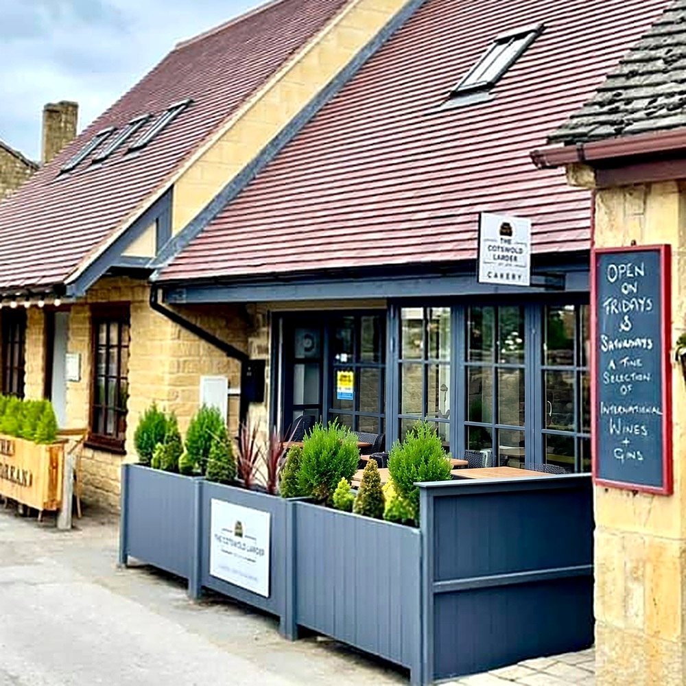 The Cotswold Larder Cakery on Kennel Lane: a honey-stone building with a red tiled roof, grey planters with small conifers, outdoor tables and a chalkboard by the door